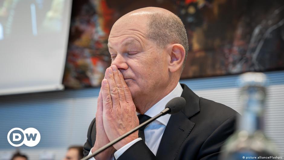 A tilted, close aerial photograph of a fighter jet flying over the sea, with land in the distance.